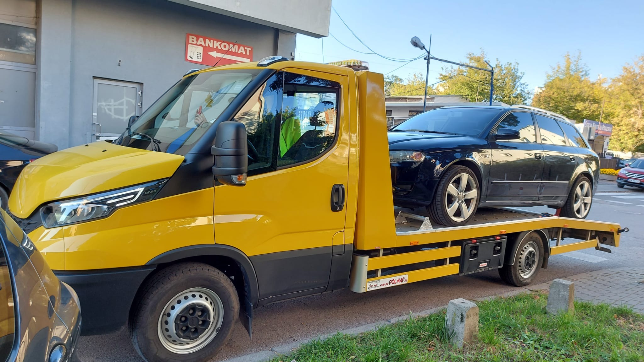 Żółta laweta Iveco z czarnym Audi A4 Avant na platformie, zaparkowana na ulicy przed budynkiem z szyldem 'Bankomat'.