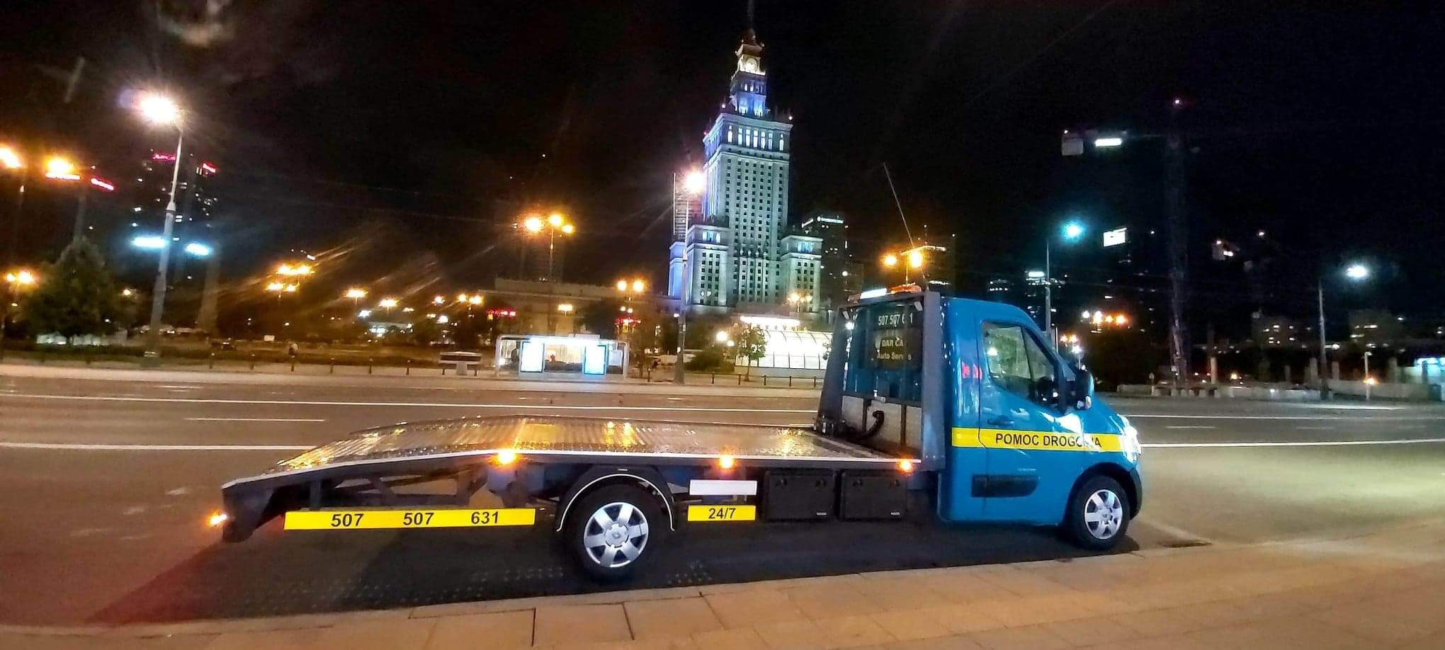 Niebieska laweta z napisem 'Pomoc Drogowa' stoi na tle nocnego krajobrazu Warszawy z Pałacem Kultury i Nauki w tle.