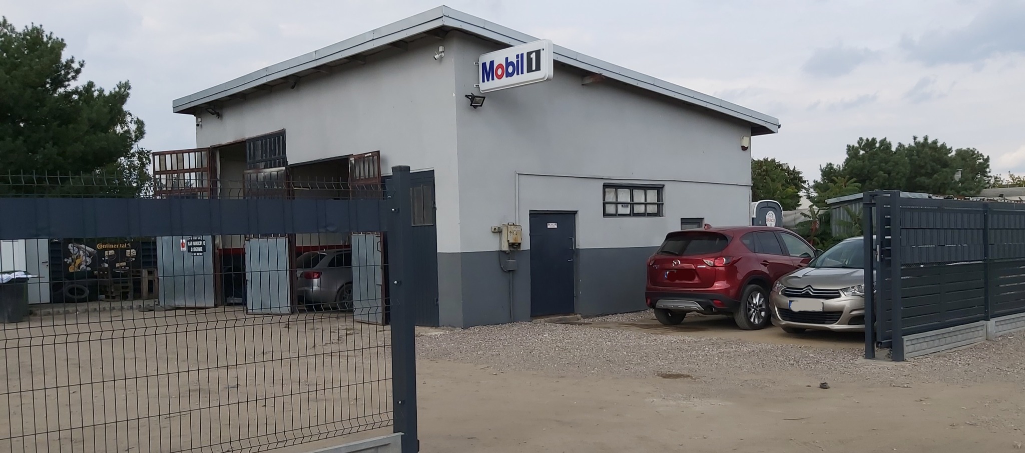 Zewnętrzny widok warsztatu samochodowego z logo Mobil 1, zaparkowanymi samochodami osobowymi i otwartymi drzwiami garażowymi, przez które widać wnętrze.