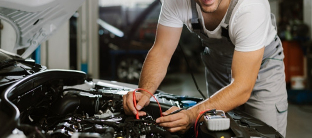 Diagnostyka silnika samochodowego: mężczyzna w kombinezonie roboczym używa multimetru do sprawdzenia instalacji elektrycznej w komorze silnika z otwartą maską.