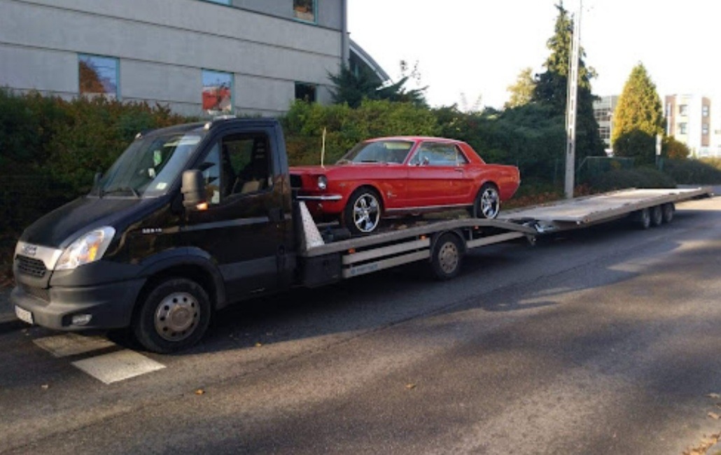 Czerwony Ford Mustang transportowany na lawecie przez czarny samochód pomocy drogowej, widok z boku na tle budynku i zieleni.