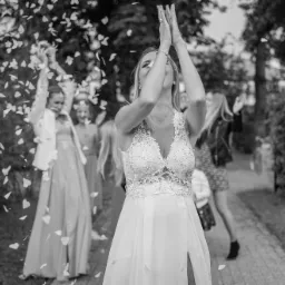 Czarno-białe zdjęcie panny młodej w koronkowej sukni, z rękami uniesionymi w górę, otoczonej przez konfetti w kształcie serc i uśmiechnięte druhny w tle.