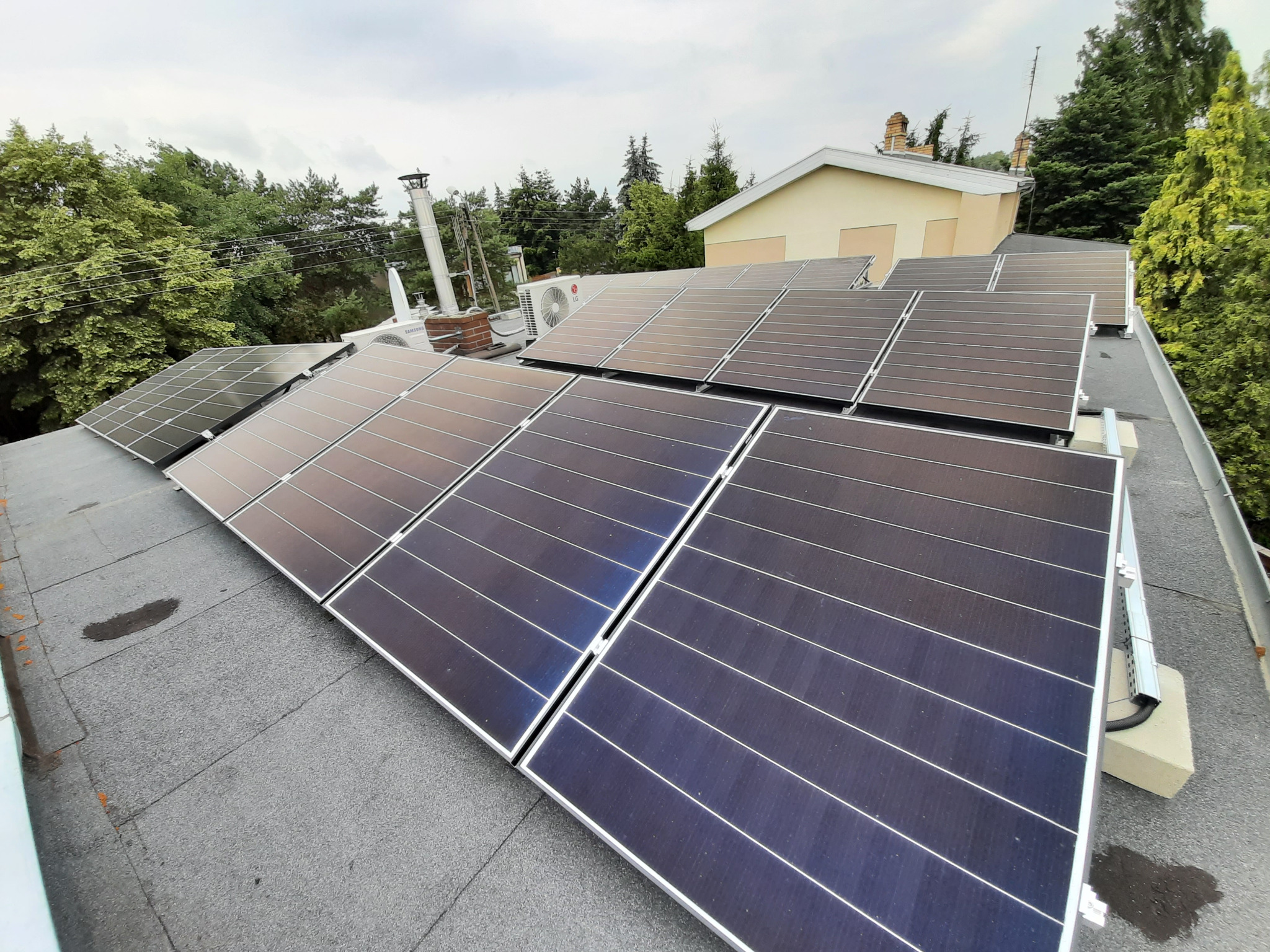 Panele słoneczne na płaskim dachu domu z widocznym kominem i klimatyzatorem, otoczone zielenią drzew.