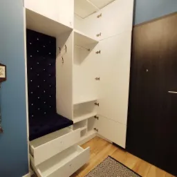 Biała szafa wnękowa z siedziskiem obitym granatowym welurem zdobionym kryształkami, otwarte szuflady z kryształowymi gałkami, drewniana podłoga z dywanikiem w geometryczne wzory, ciemne drzwi...