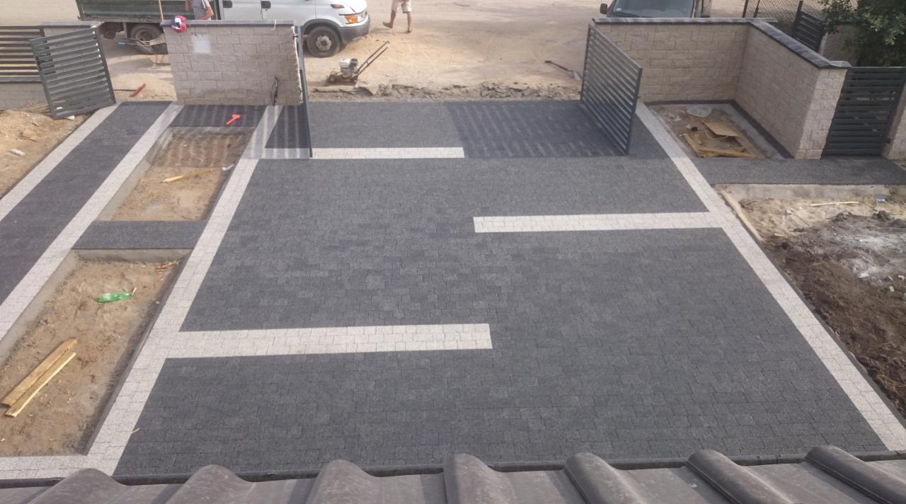 Ukończony podjazd z grafitowej kostki brukowej z jasnymi pasami, obrzeża i przygotowane rabaty. W tle widoczny samochód dostawczy i ogrodzenie.