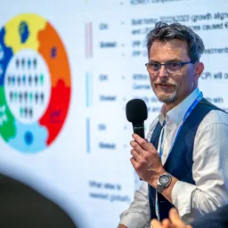 Prelegent z mikrofonem podczas konferencji TÜV SÜD, na ekranie za nim prezentacja z kolorowym schematem kołowym i tekstem.