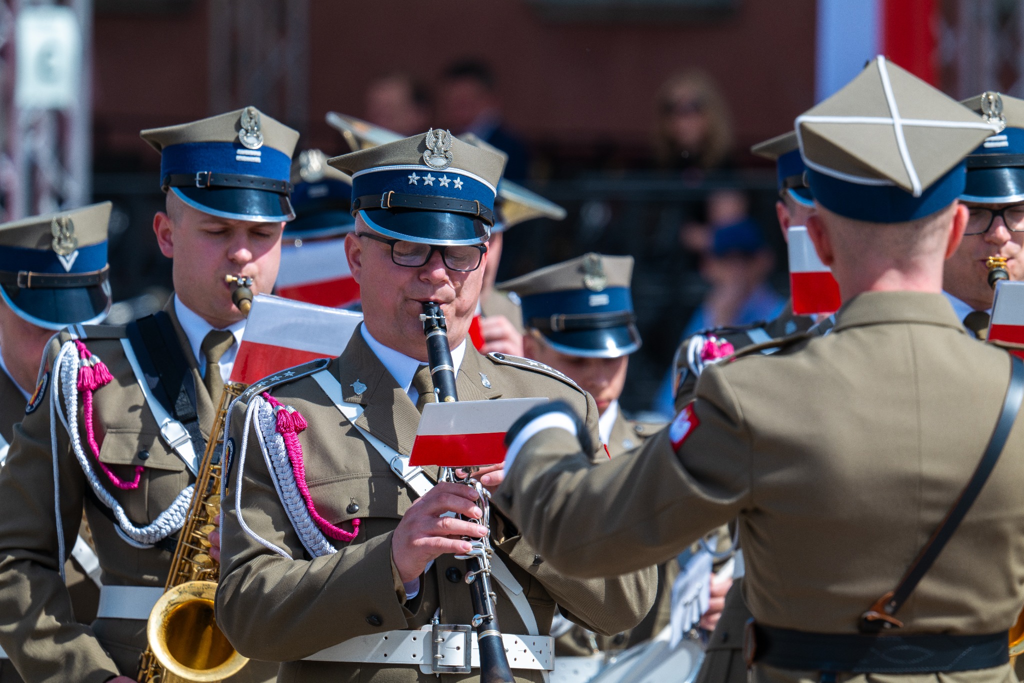 Orkiestra wojskowa w mundurach grająca na instrumentach dętych podczas obchodów 3 Maja na Placu Zamkowym w Warszawie. Widoczny klarnecista w okularach i saksofonista z ozdobnymi sznurami.