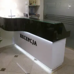 Nowoczesna lada recepcyjna z czarnym, falistym blatem i podświetlanym napisem 'RECEPCJA', uzupełniona rośliną doniczkową i dekoracyjną szybą z motywem muzycznym.