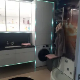 Nowoczesna łazienka z podświetlanymi szafkami, umywalką z czarnym blatem, pralką w zabudowie i wanną z hydromasażem za szklaną ścianką.