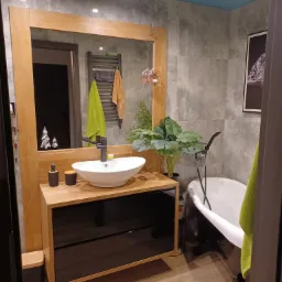 Nowoczesna łazienka z umywalką nablatową w kształcie misy, szafką z czarnymi frontami i drewnianym blatem, oraz wolnostojącą wanną na lwich łapach, uzupełniona o rośliny doniczkowe i szare płytki...