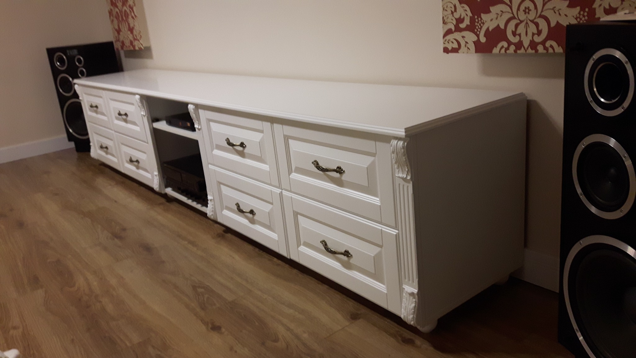 Długa, biała komoda RTV z frezowanymi zdobieniami i metalowymi uchwytami szuflad, stojąca na drewnianej podłodze, obok czarnych głośników.