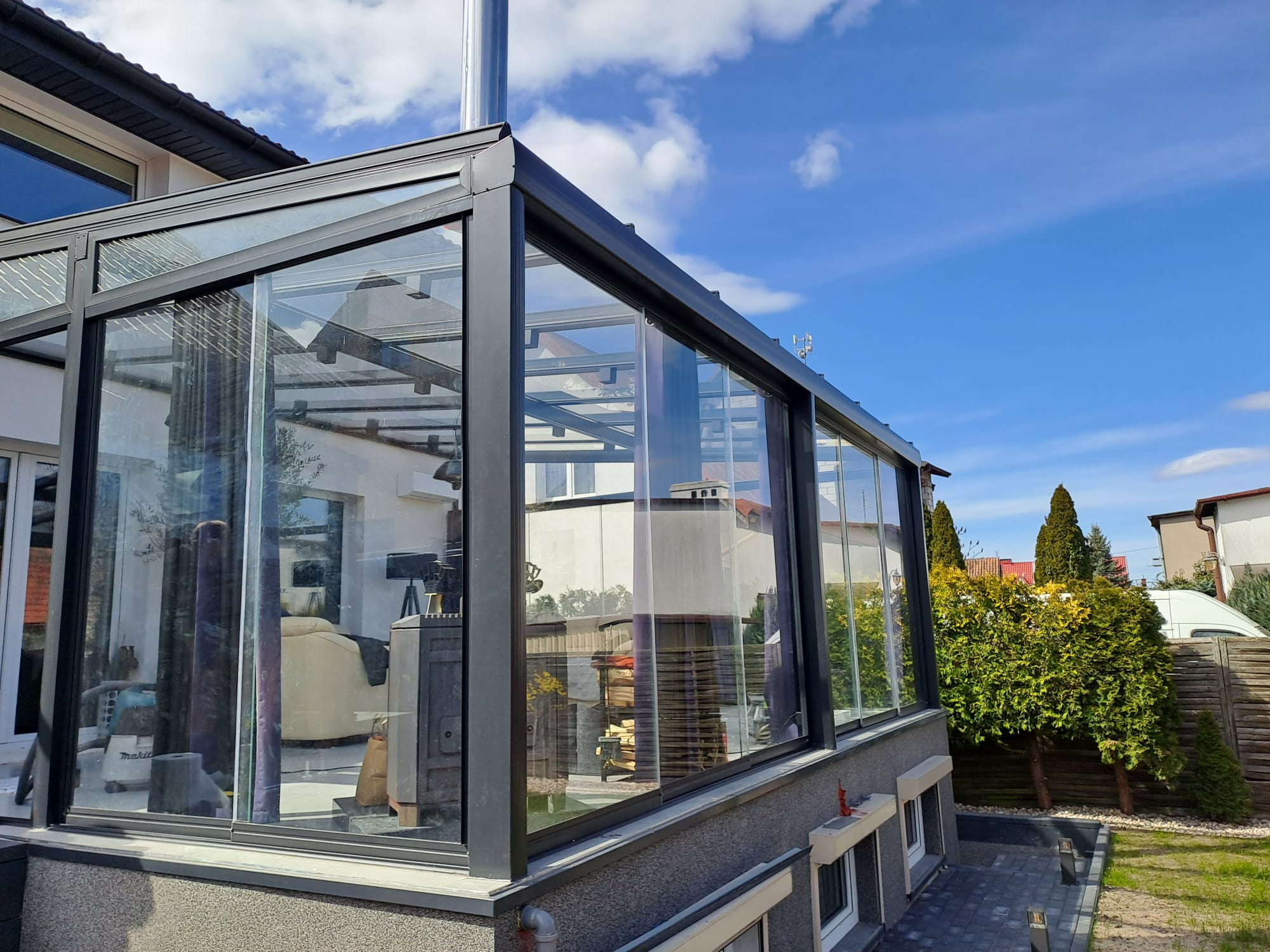 Szklana zabudowa tarasu z czarną ramą, widok z zewnątrz. Wnętrze tarasu z kominkiem i meblami. Błękitne niebo w tle. Detal: widoczny komin.