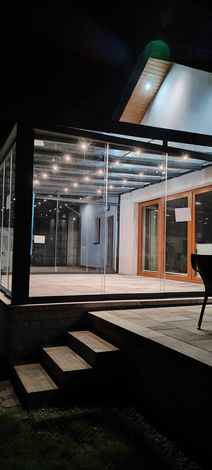 Zabudowany taras z przeszklonymi ścianami i dachem, oświetlony girlandą żarówek. Widoczne schody wejściowe i fragment krzesła. Nocne ujęcie.