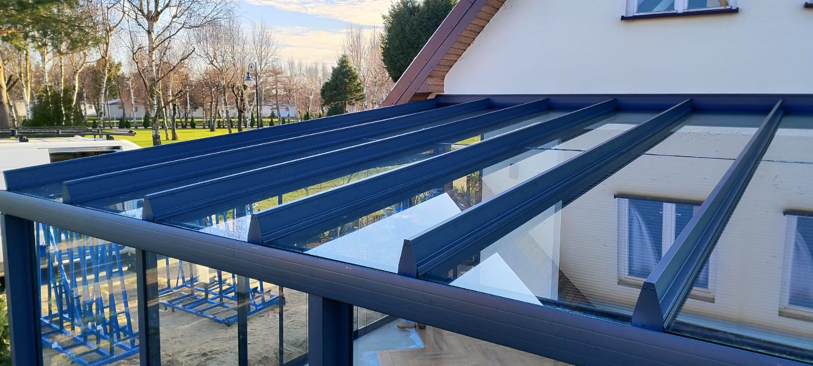 Zadaszenie tarasu z niebieską konstrukcją i szklanym wypełnieniem, widok z dołu, z odbiciem otoczenia w szkle. Nowoczesny design, jasne otoczenie.