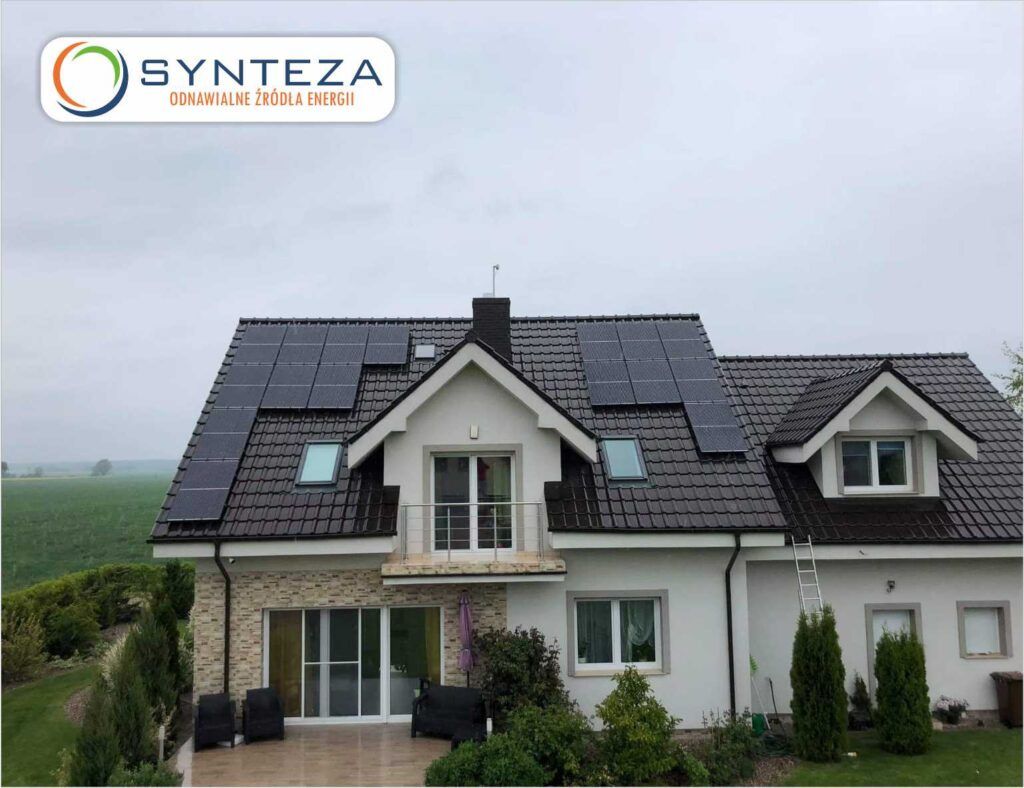 Dom jednorodzinny z ciemną dachówką i panelami słonecznymi na dachu, widok z zewnątrz w pochmurny dzień. Na szczycie dachu widoczny maszt antenowy.