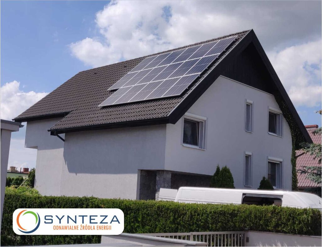 Dwuspadowy dom z szarymi panelami słonecznymi na dachu pokrytym brązową dachówką, biała elewacja, logo firmy Synteza na pierwszym planie.