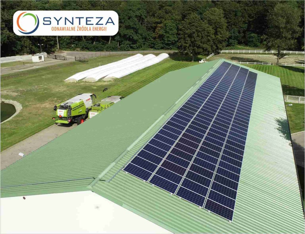 Zielony dach budynku gospodarczego pokryty panelami fotowoltaicznymi; w tle kombajn rolniczy i białe tunele foliowe.