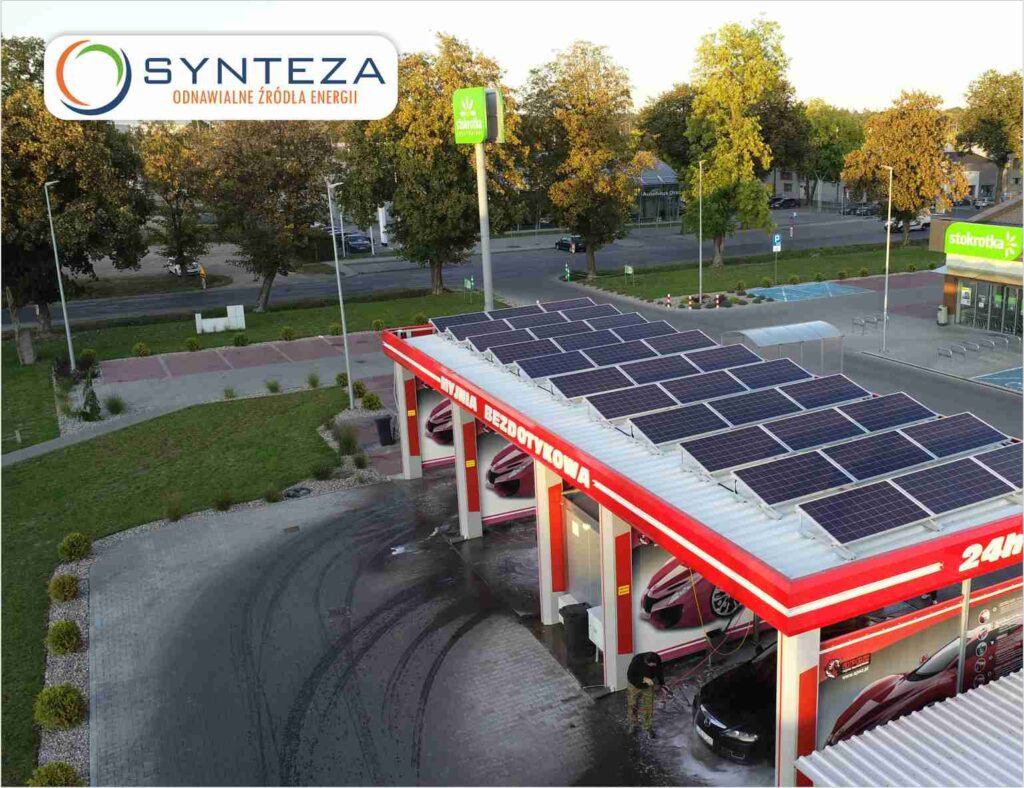 Myjnia samochodowa z panelami słonecznymi na dachu, widok z góry, pracownik obsługujący czarny samochód. W tle budynek Stokrotki.