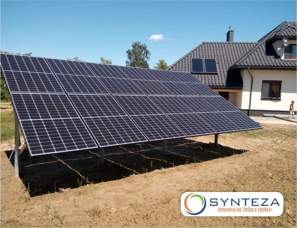 Panele słoneczne na metalowej konstrukcji przed domem jednorodzinnym z dodatkowymi panelami na dachu, widoczne logo firmy w prawym dolnym rogu.