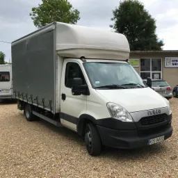 Biały samochód dostawczy Iveco z szarą plandeką na tle szarego nieba i budynku z reklamą okien, zaparkowany na żwirowym podłożu.