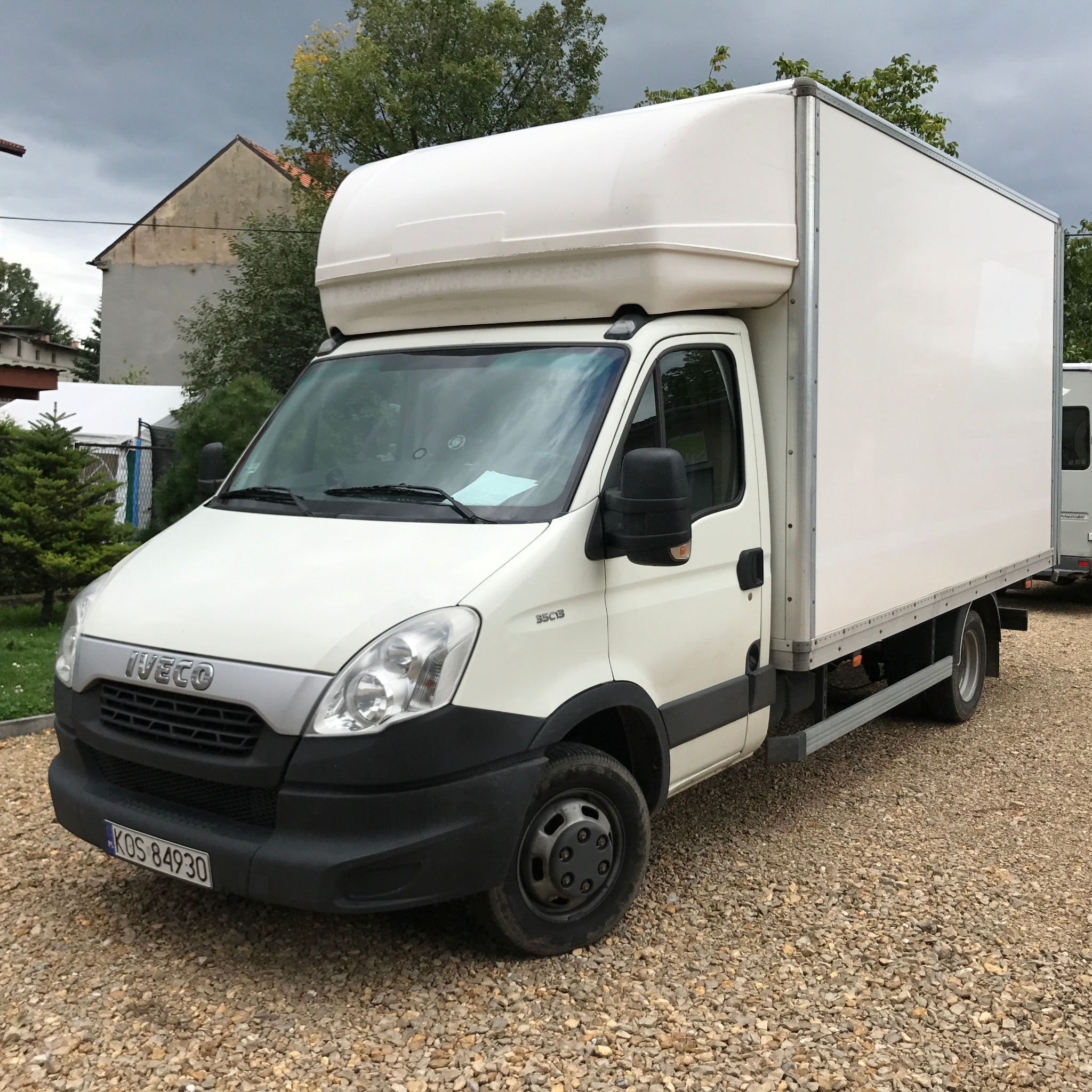 Biały samochód dostawczy Iveco z wysoką zabudową na żwirowym podłożu, tablica rejestracyjna KOS 84930, pochmurne niebo w tle.