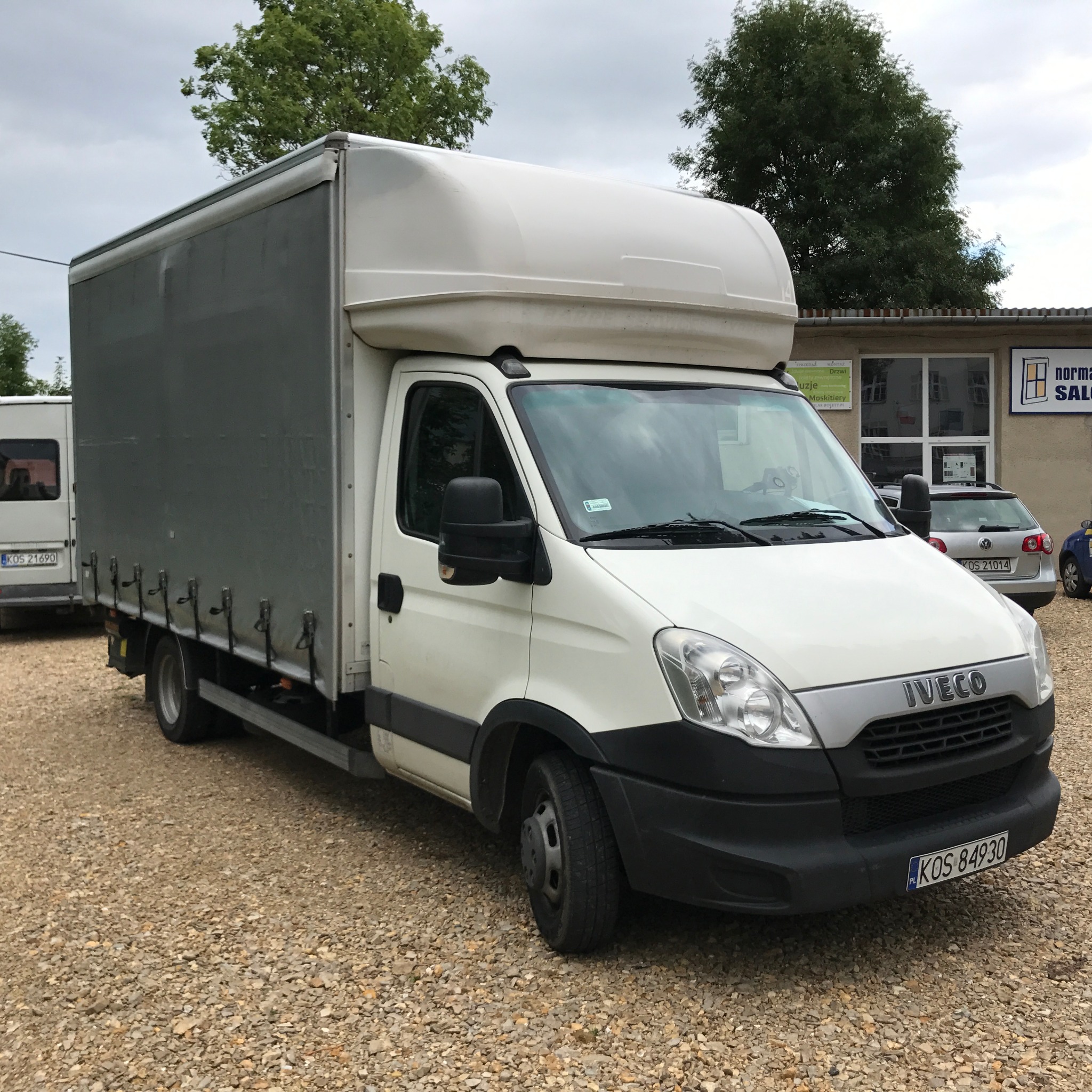 Biały samochód dostawczy Iveco z szarą plandeką na tle szarego nieba i budynku z reklamą okien, zaparkowany na żwirowym podłożu.