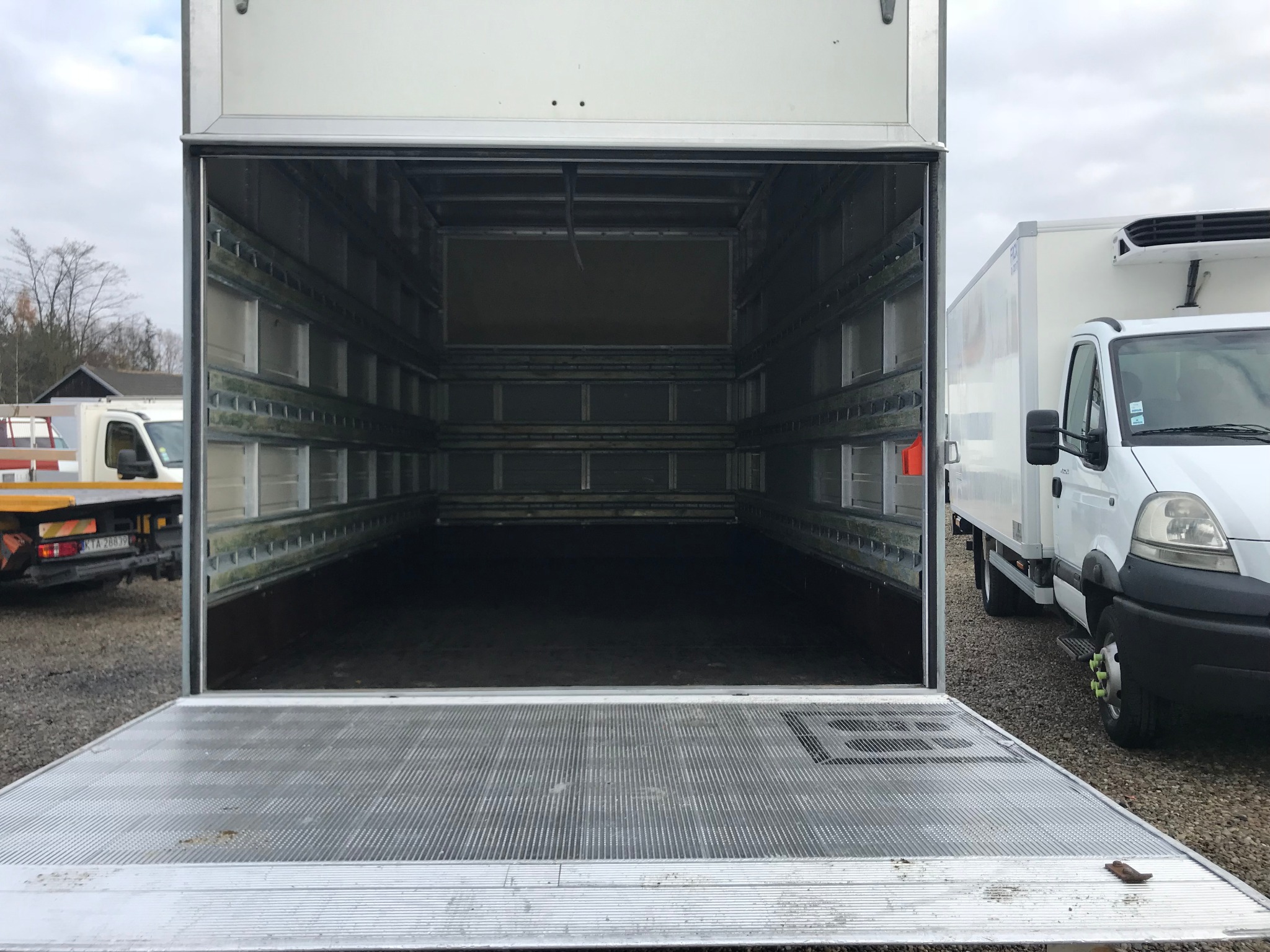 Otwarta pusta przestrzeń ładunkowa chłodni z widocznymi półkami, na zewnątrz widoczne inne pojazdy dostawcze na żwirowym podłożu.