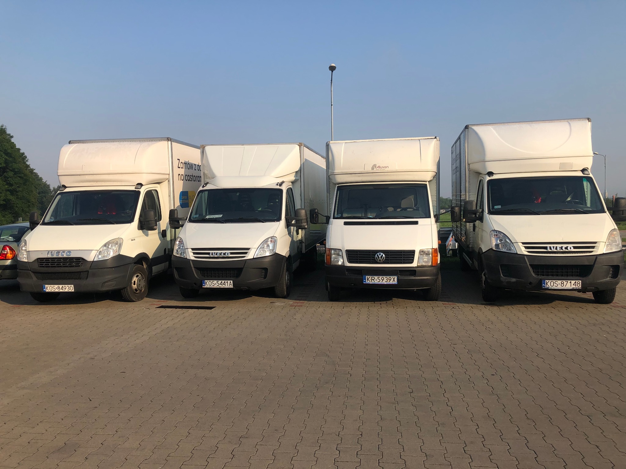 Cztery białe busy dostawcze różnych marek ustawione w rzędzie na brukowanym parkingu w słoneczny dzień, widoczne numery rejestracyjne z różnych miast.