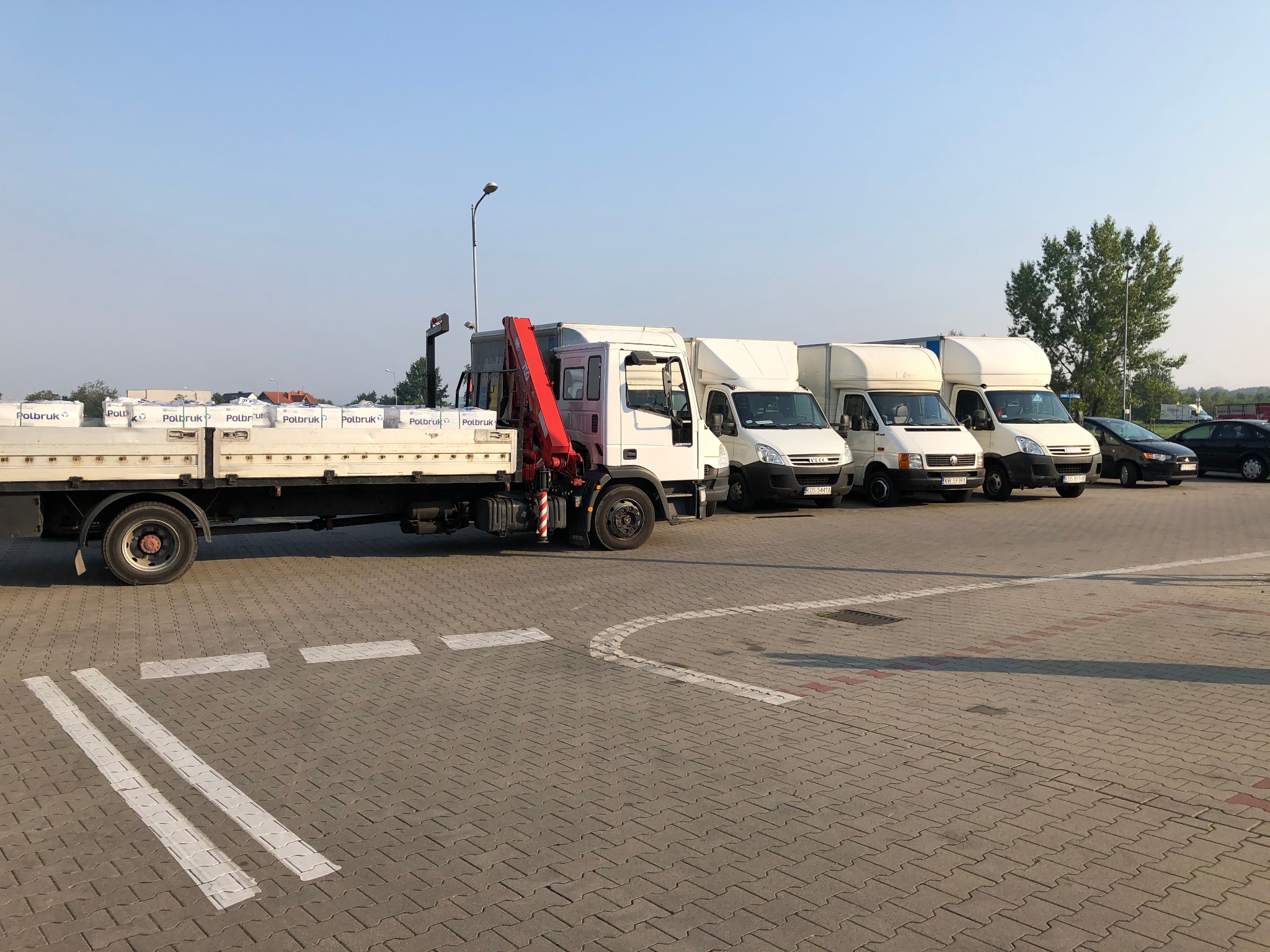 Ciężarówka z czerwoną dźwignią załadowana kostką brukową Polbruk, zaparkowana obok trzech busów na parkingu z kostki brukowej.