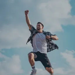 Mężczyzna w skoku, z podniesioną pięścią, wyrażający entuzjazm na tle błękitnego nieba z chmurami, nad nim logo 'Empowerment Coaching' i hasło 'Be a voice, not en echo'.