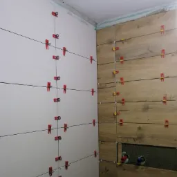 Ściana w trakcie układania płytek, widoczne płytki drewnopodobne i białe, system poziomowania z czerwonymi i żółtymi klinami, otwory na rury hydrauliczne.