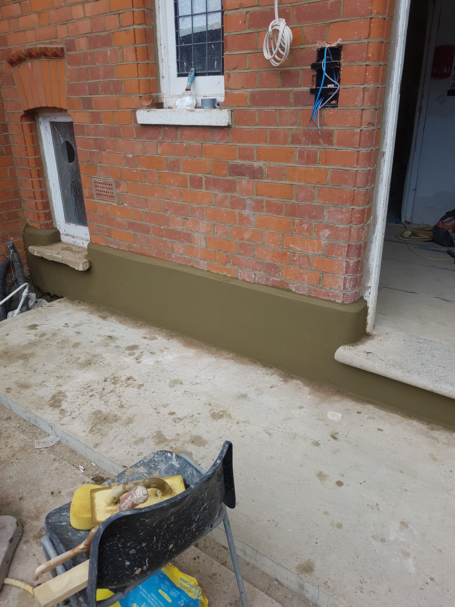 Świeżo otynkowana dolna część elewacji z czerwonej cegły przy wejściu do budynku, widoczne narzędzia w wiadrze i wystające przewody elektryczne.