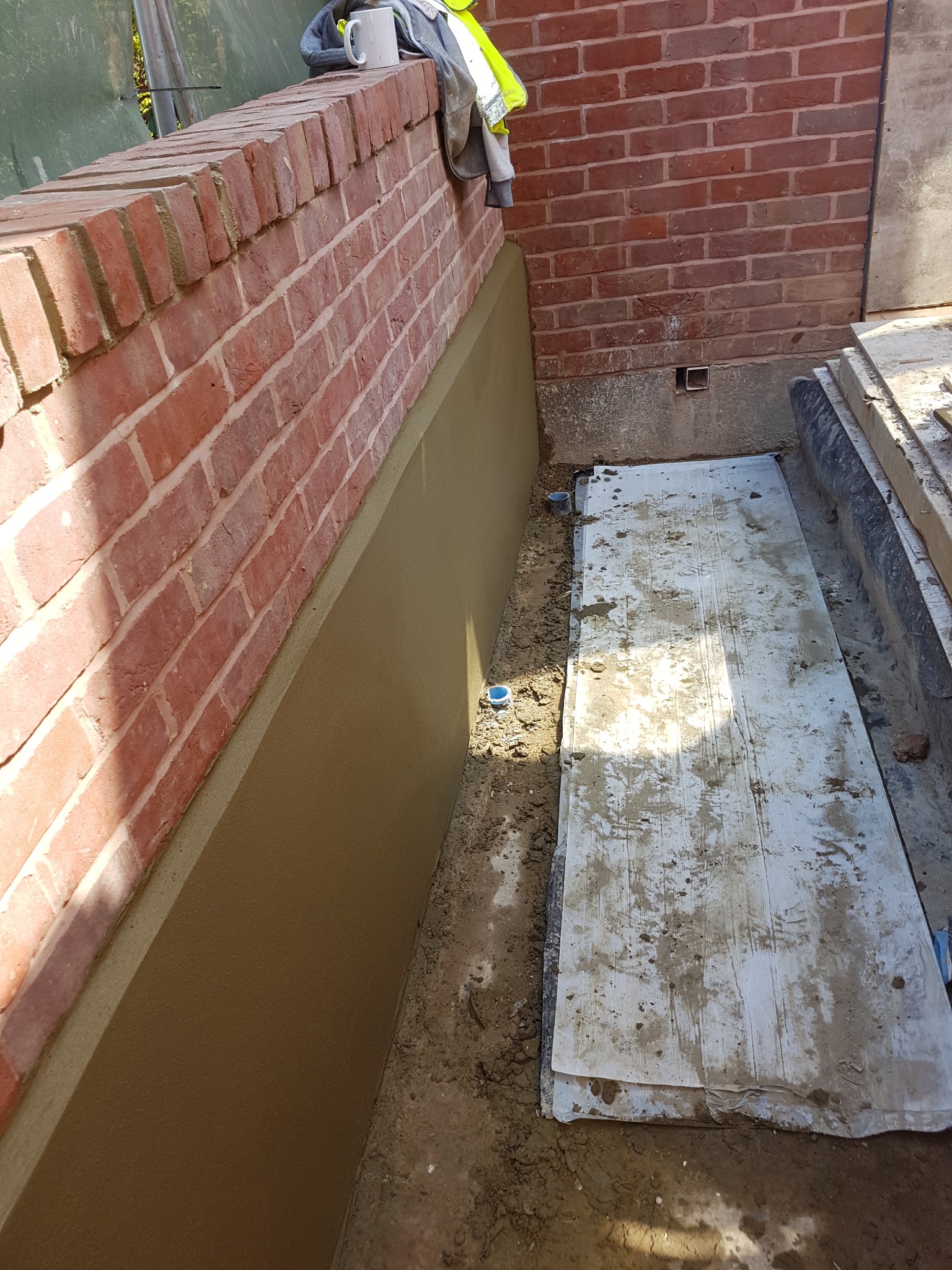 Ceglany murek z otynkowaną dolną częścią, obok ziemia i płyty budowlane, na murku kubek i kamizelka odblaskowa.