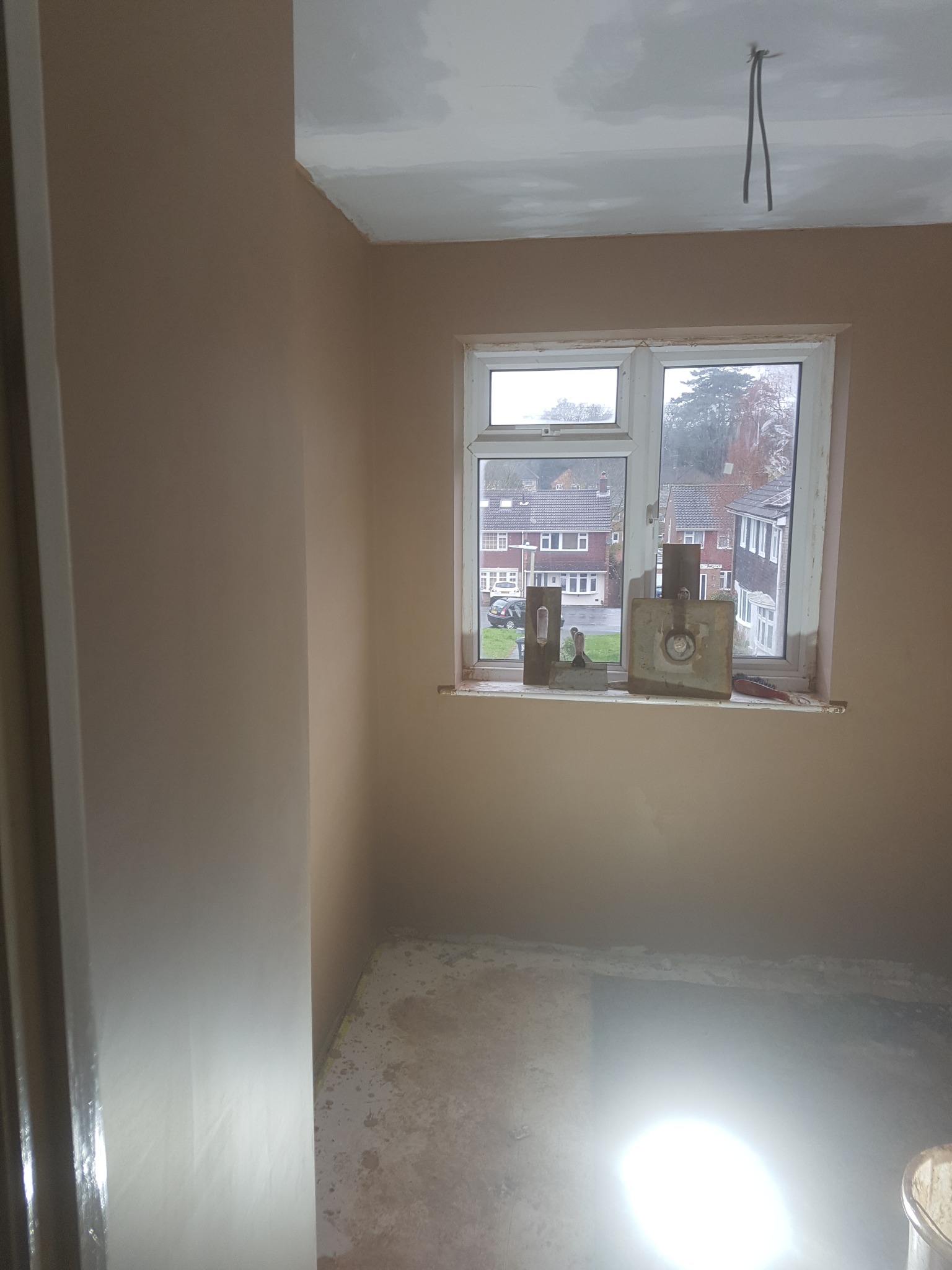 Pokój w trakcie prac wykończeniowych: ściany pokryte świeżym tynkiem, narzędzia na parapecie okna z widokiem na domy w tle, widoczne kable wystające z sufitu.