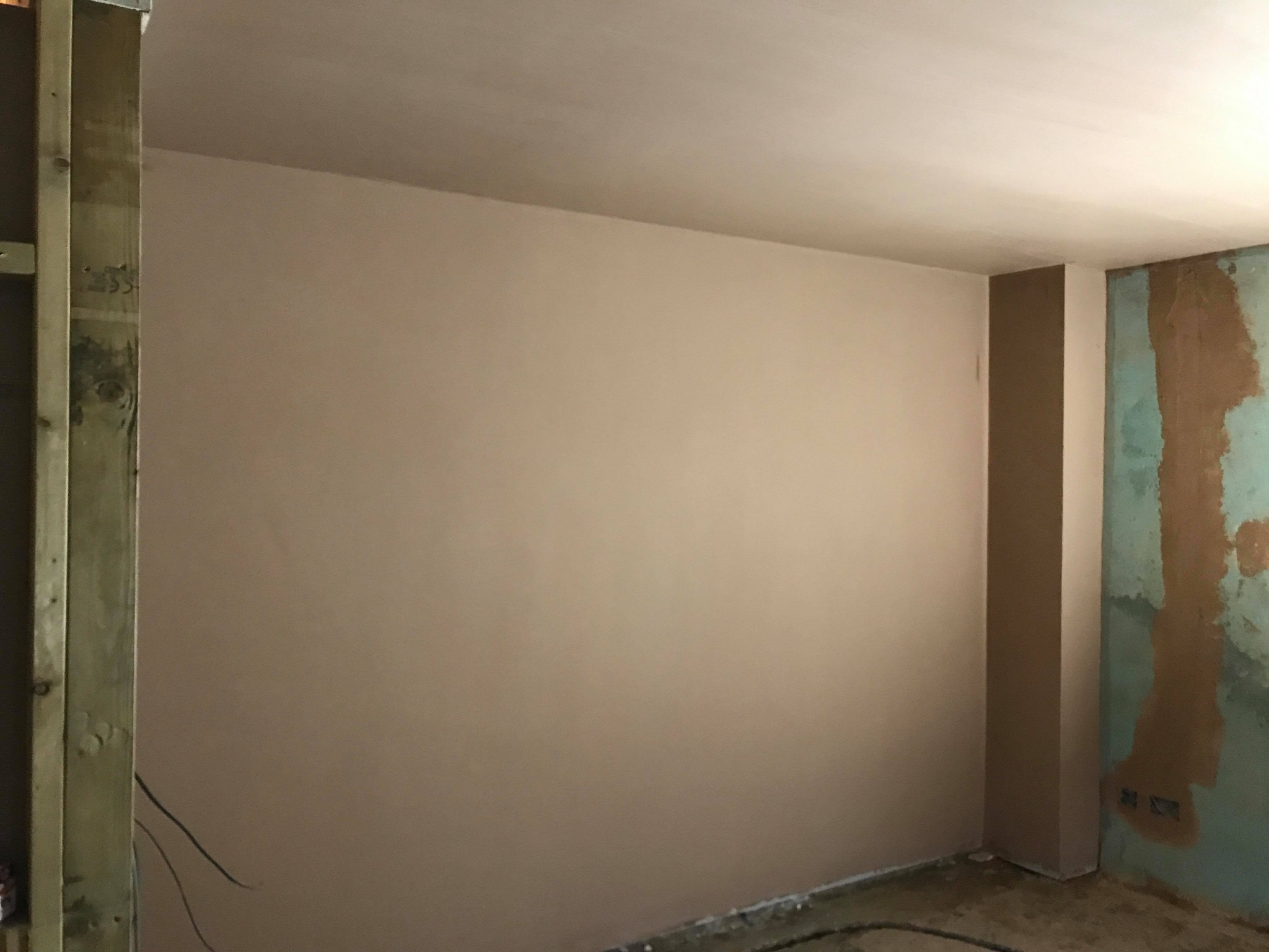 Świeżo otynkowana ściana w pomieszczeniu w trakcie remontu, widoczne drewniane elementy konstrukcyjne i fragment starej ściany z pozostałościami farby.