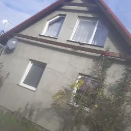 Ocieplony dom jednorodzinny z szarą elewacją, widoczne okna z białymi ramami, brązowe elementy drewniane pod dachem i satelita na ścianie, na belce siedzi czarny ptak