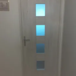 Białe drzwi wewnętrzne z trzema prostokątnymi przeszkleniami, srebrna klamka, widoczny fragment obrazu na ścianie.