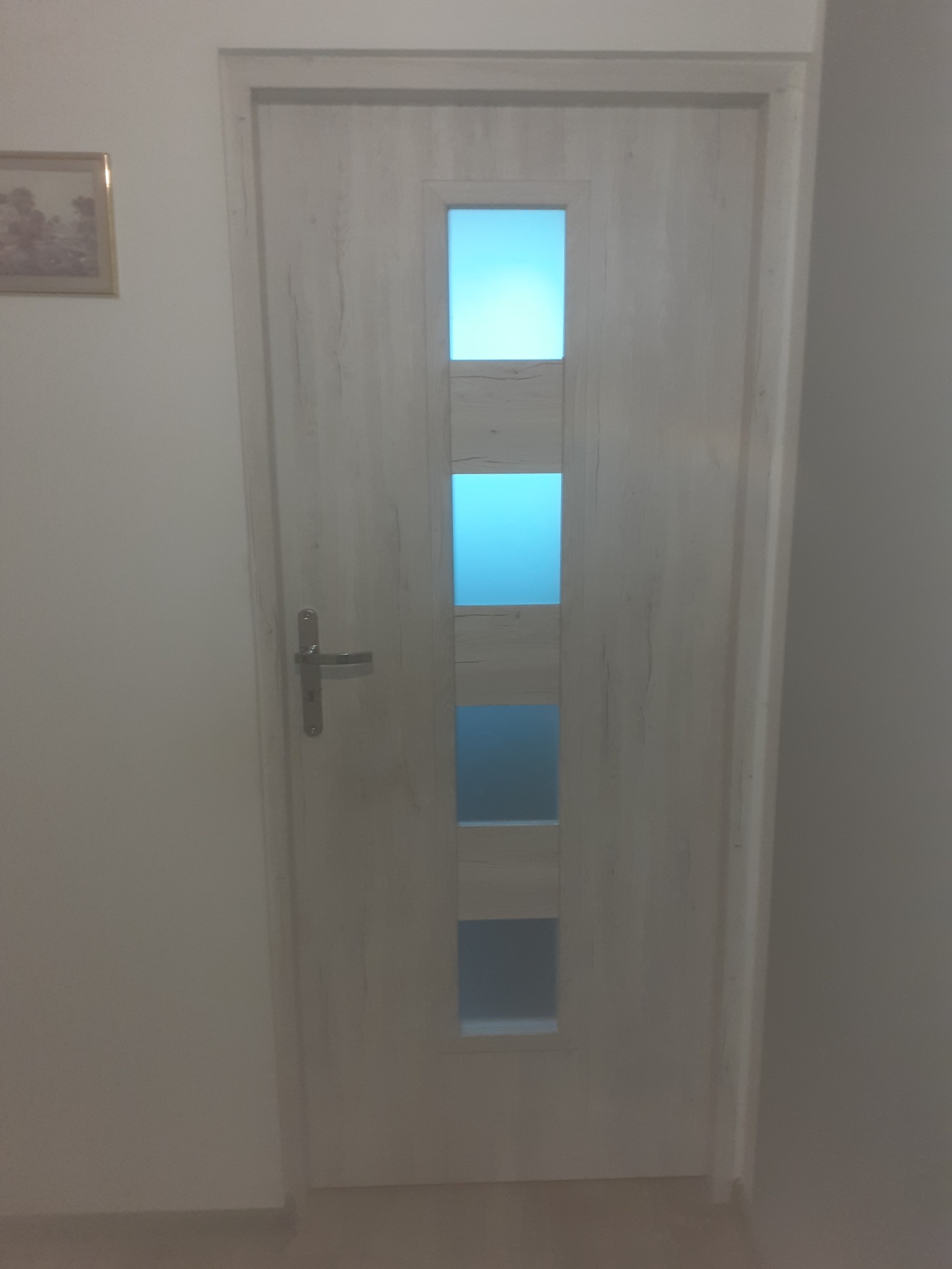 Białe drzwi wewnętrzne z trzema prostokątnymi przeszkleniami, srebrna klamka, widoczny fragment obrazu na ścianie.