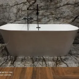 Wolnostojąca wanna w nowoczesnej łazience z marmurowymi płytkami na ścianach i podłodze, czarna armatura i drewnopodobne panele podłogowe w dolnej części kadru.