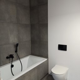 Ale remont! Monika Kwapiś-Skurzewska - Nowoczesna łazienka z szarymi płytkami, białą wanną z czarną armaturą i podwieszaną toaletą z czarnym panelem spłukującym.