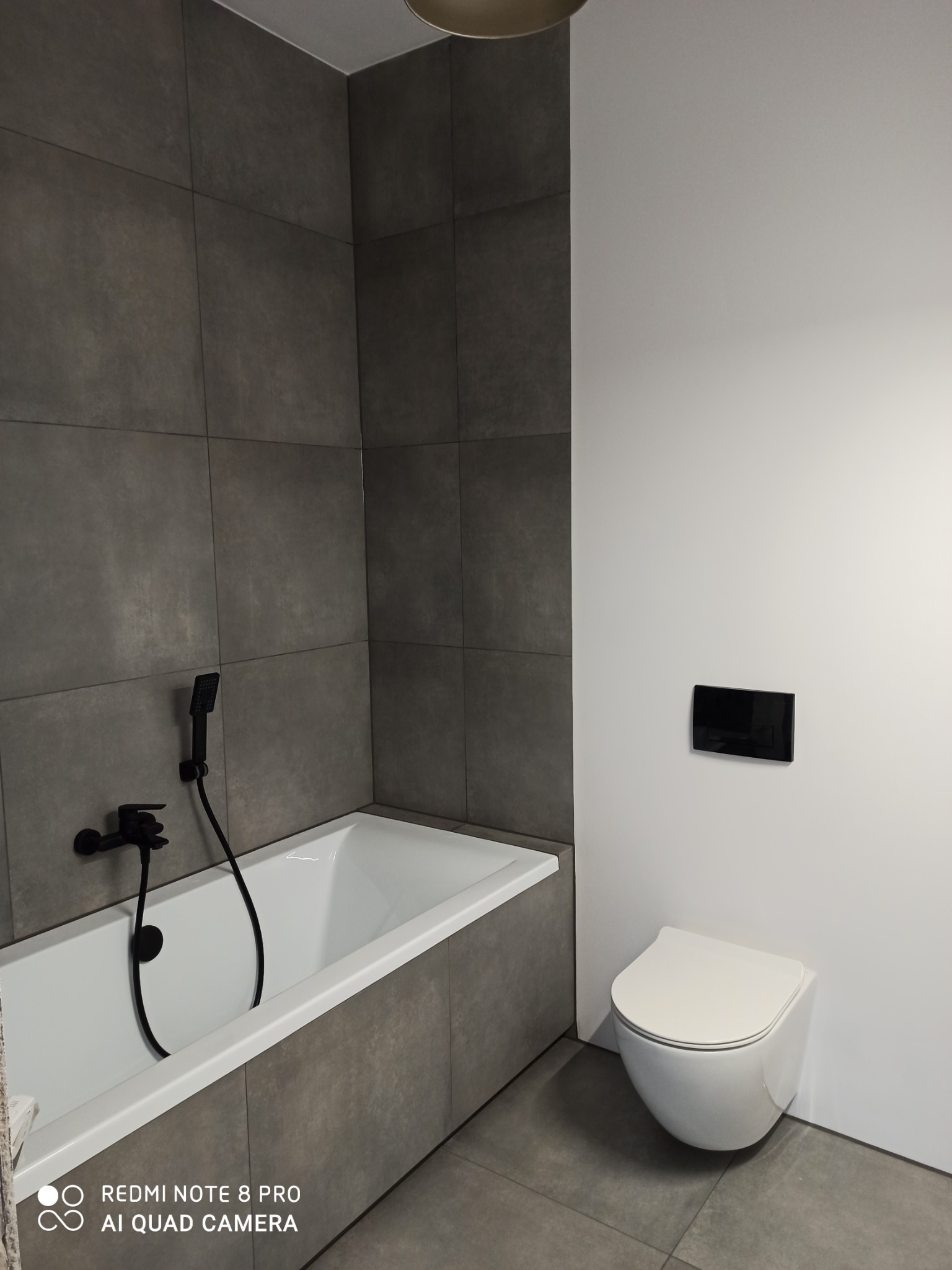 Nowoczesna łazienka z szarymi płytkami, białą wanną z czarną armaturą i podwieszaną toaletą z czarnym panelem spłukującym.