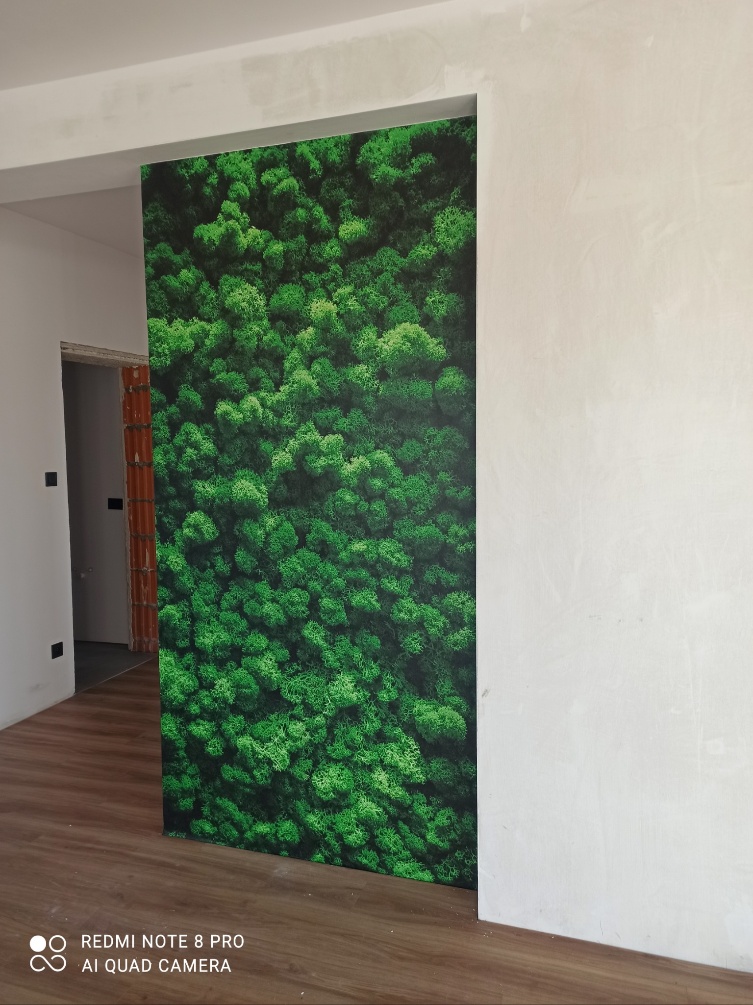 Panel dekoracyjny z motywem mchu na ścianie w surowym wnętrzu, widoczny fragment podłogi i ściany w stanie deweloperskim.