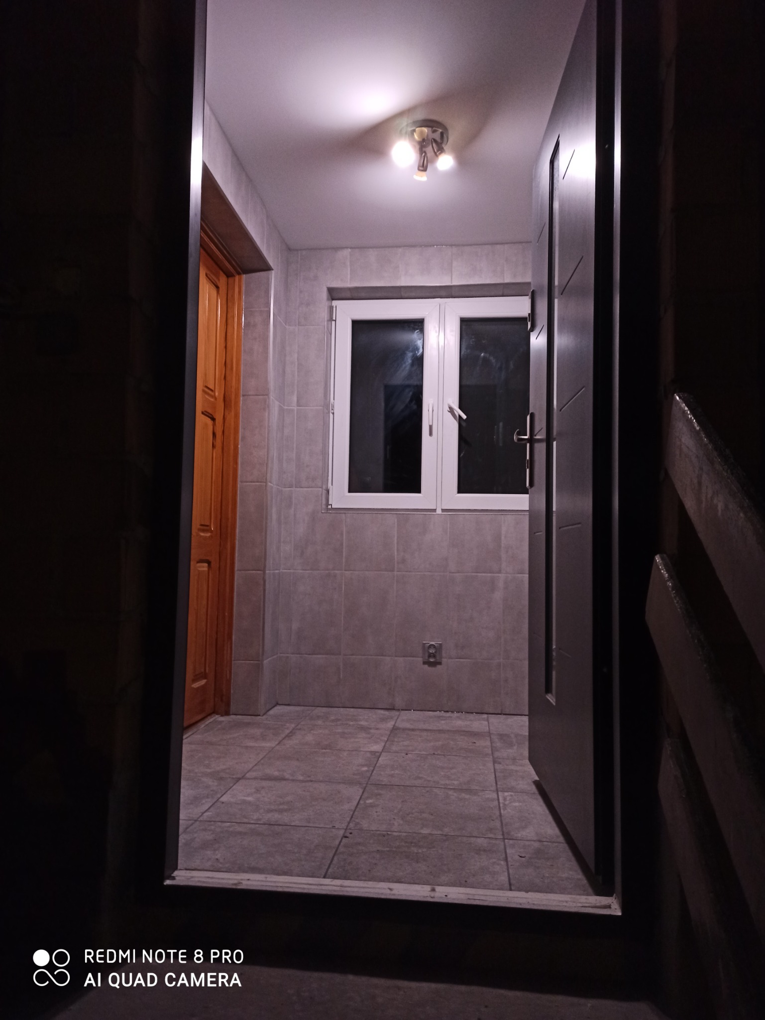 Wyremontowana łazienka z szarymi płytkami, białym oknem i otwartymi drzwiami wejściowymi, widok z zewnątrz.