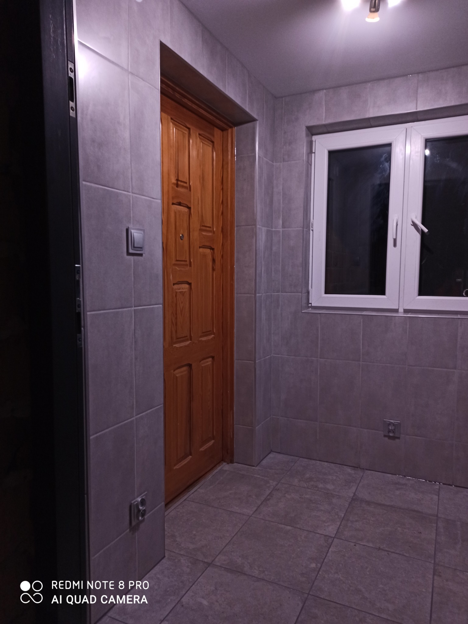 Wnętrze łazienki w Nieporęcie po remoncie: szare płytki na ścianach i podłodze, drewniane drzwi wejściowe, białe okno z widokiem na zewnątrz.