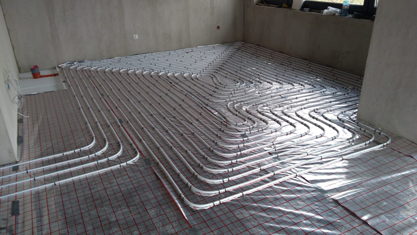 Montaż ogrzewania podłogowego: białe rurki ułożone w pętle na folii z czerwoną siatką w surowym pomieszczeniu przed wylewką.