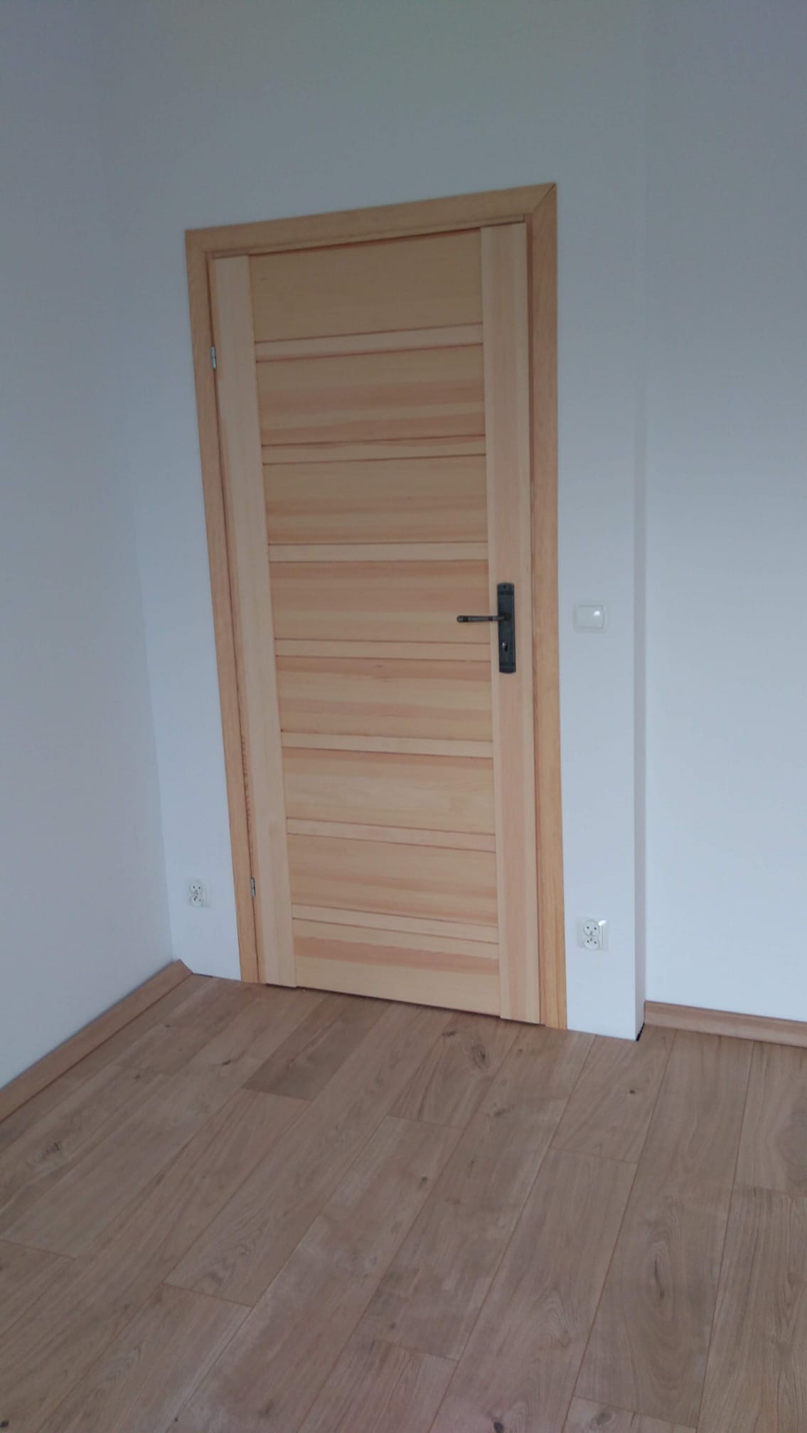 Zamknięte drzwi wewnętrzne z drewna o jasnym odcieniu, z poziomymi panelami, czarną klamką i ościeżnicą z widocznymi słojami, na tle białej ściany i paneli podłogowych imitujących drewno.