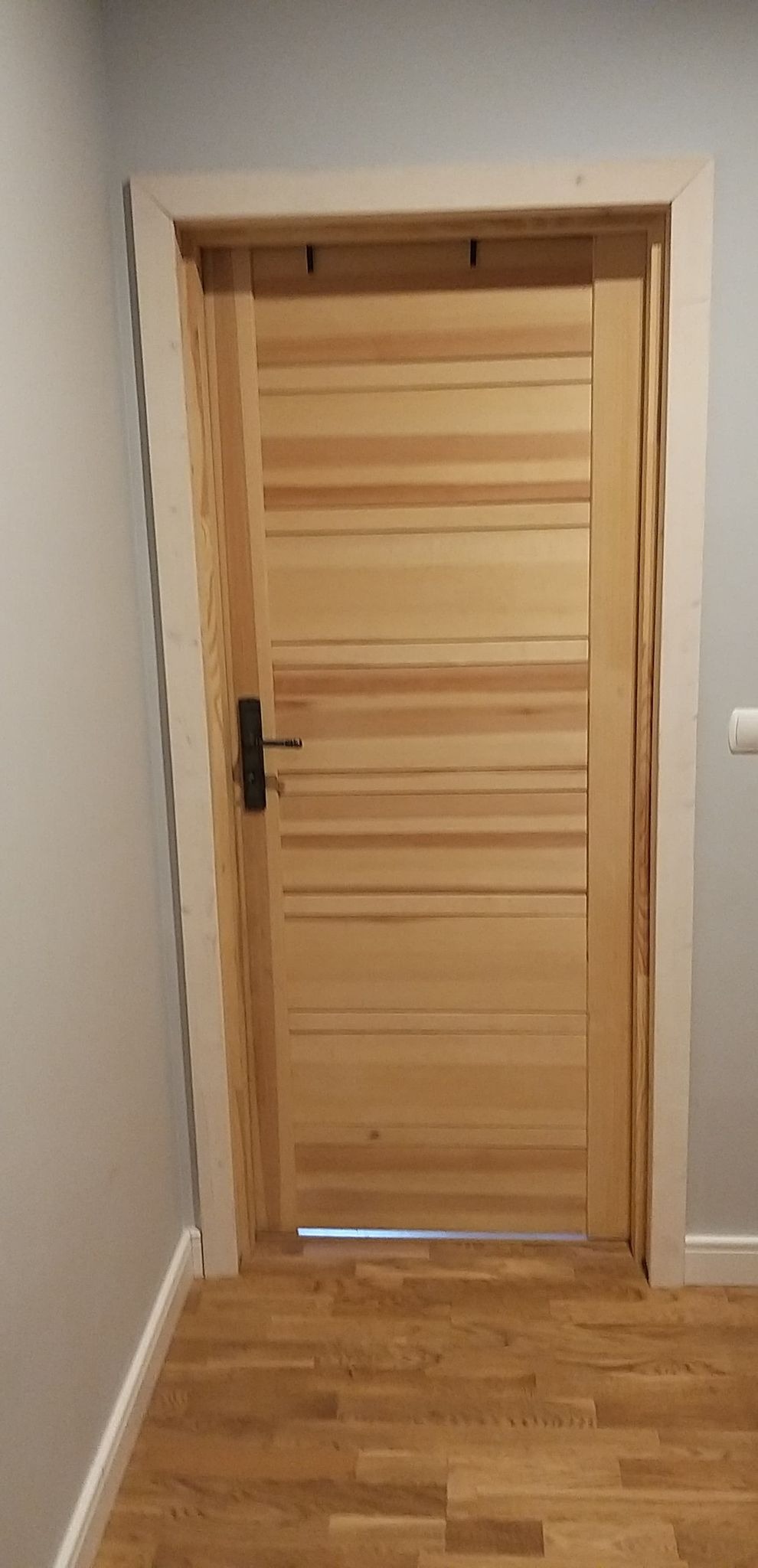 Zamknięte, drewniane drzwi wewnętrzne z poziomymi panelami, czarną klamką i ościeżnicą w kolorze złamanej bieli, zamontowane w szarej ścianie nad drewnianą podłogą z parkietu.