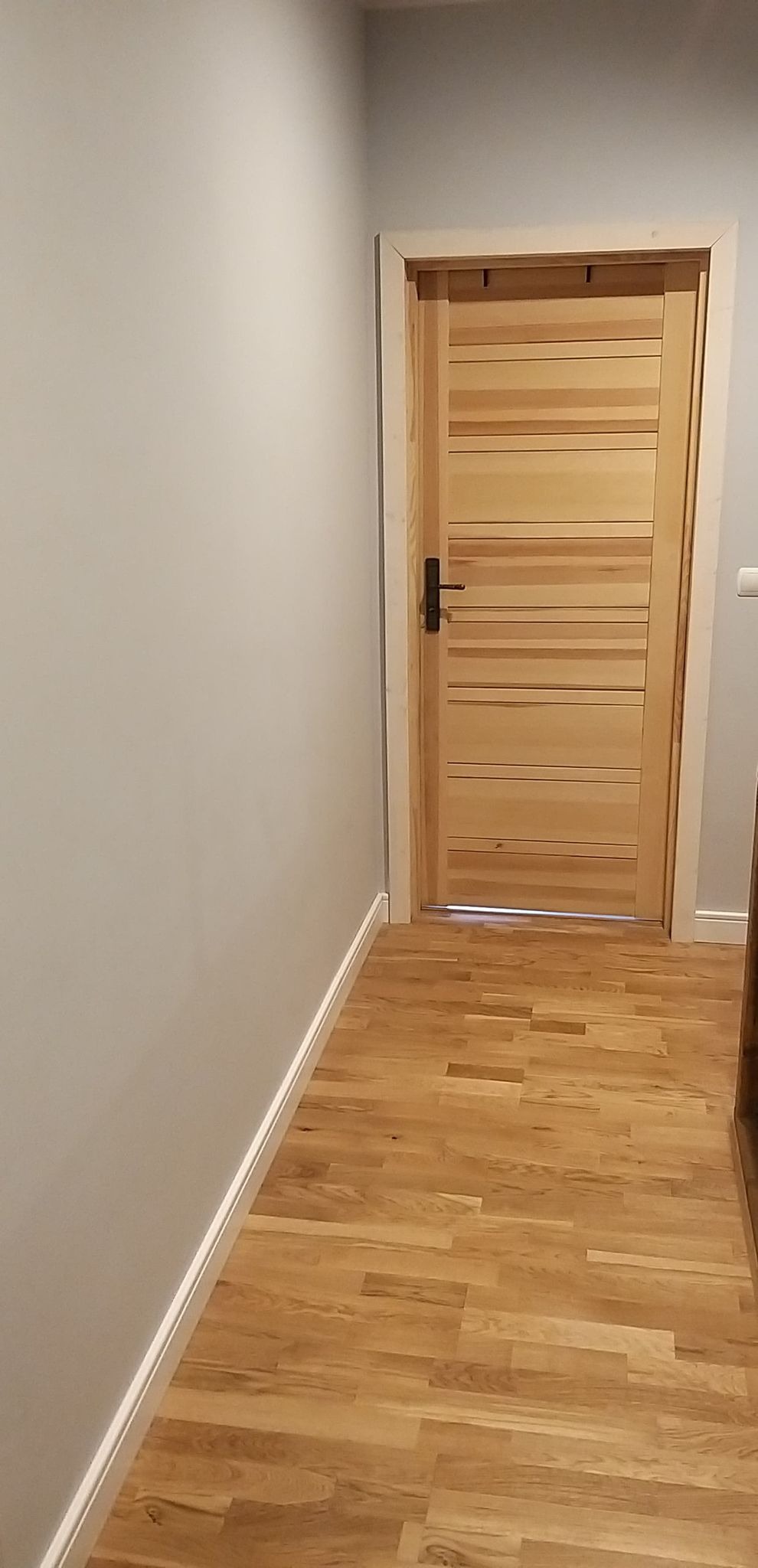 Wykończony korytarz z drewnianą podłogą, gładkimi szarymi ścianami i jasnymi drewnianymi drzwiami z poziomymi panelami i czarną klamką.