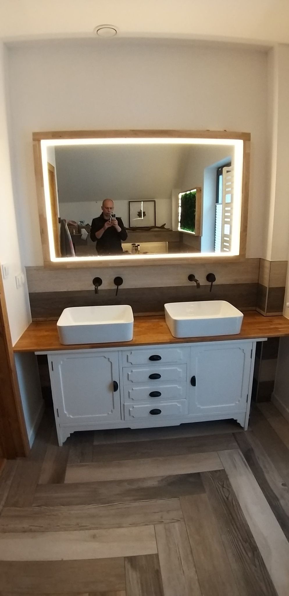 Łazienka po renowacji: biała szafka w stylu retro z dębowym blatem, dwie kwadratowe umywalki nablatowe i czarne baterie podtynkowe. Nad szafką duże lustro w dębowej ramie z podświetleniem LED,...