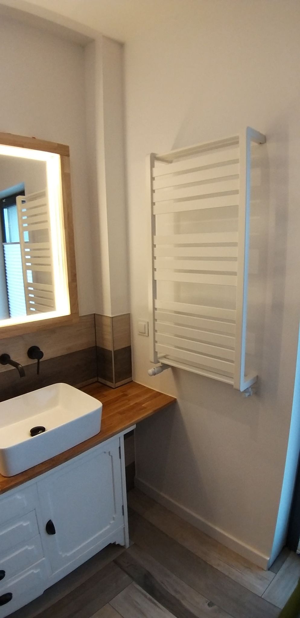 Nowoczesna łazienka z białym grzejnikiem drabinkowym, umywalką nablatową na dębowym blacie i podświetlanym lustrem z drewnianą ramą. Bateria podtynkowa w kolorze czarnym. Podłoga wyłożona płytkami...